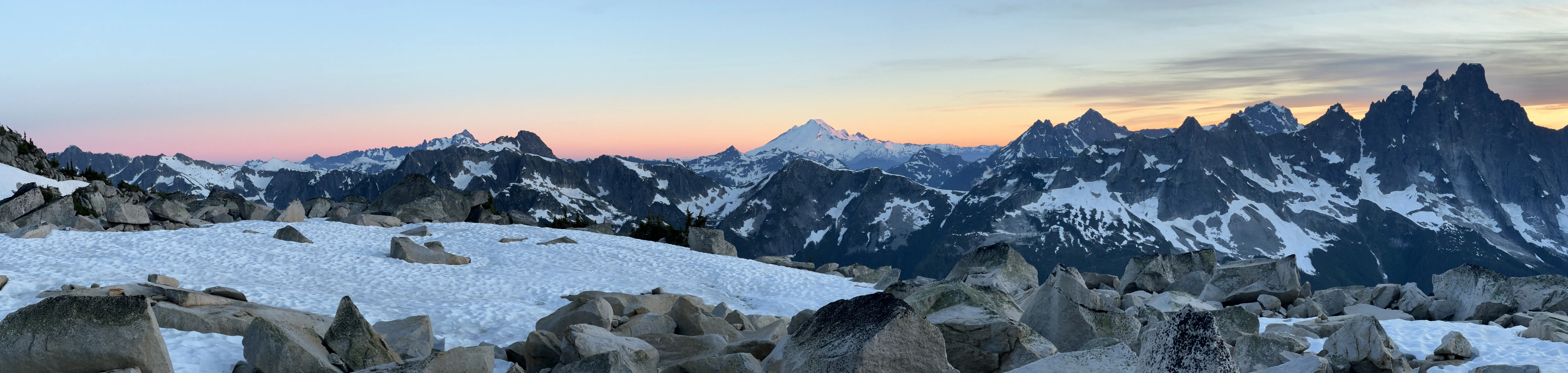 A snow-covered alpine basin below a long skyline of dark, jagged peaks at sunrise